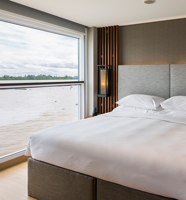 A cosy bedroom in a Deluxe Suite on the Scenic Spirit ship, with a comfortable Queen bed next to a large window, offering views of the passing river.