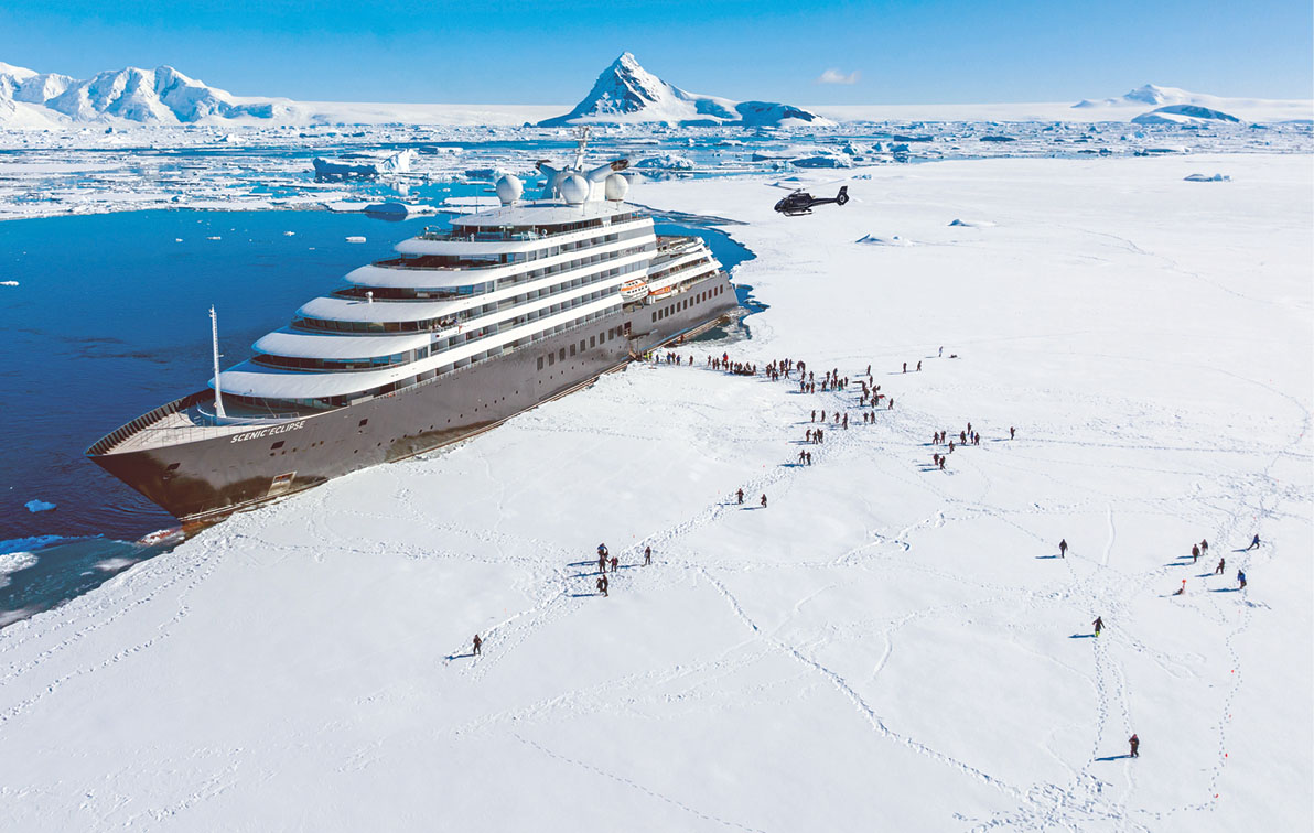 Luxury cruise ship docked on the edge of an ice sheet in Antarctica with groups of people leaving the ship to explore on the ice under sunny blue skies with a helicopter overhead.
