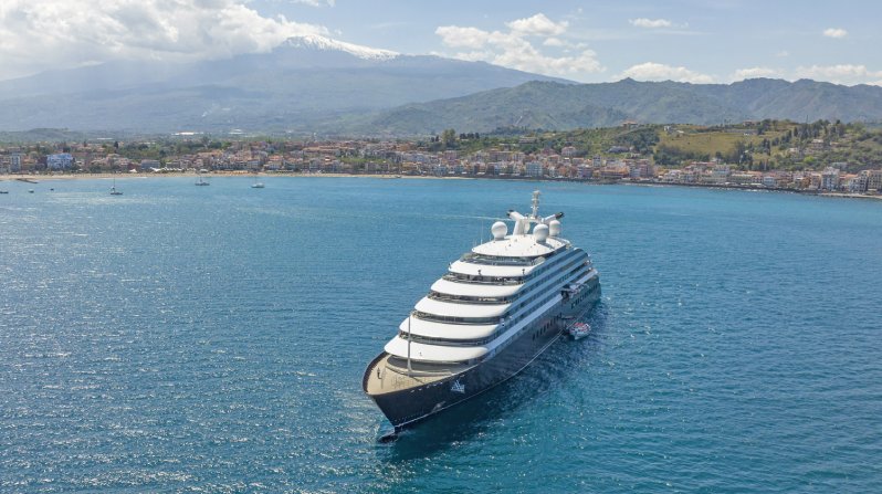 Exterior view of the Scenic Eclipse yacht near a shoreline with mountains in the distance